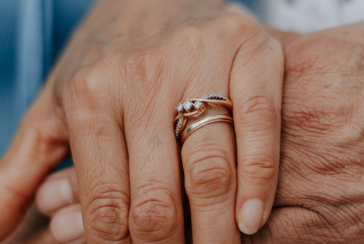 A close up of two aged hands holding each other, showcasing delicate nature-inspired rose gold wedding bands on a finger.
