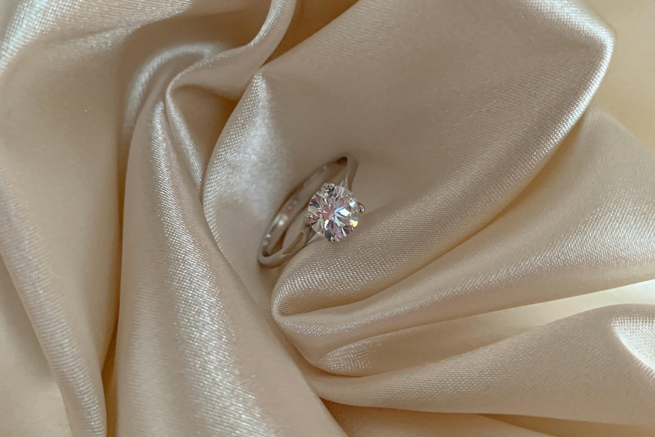 A sparkling solitaire diamond engagement ring rests on smooth, cream-colored satin fabric.